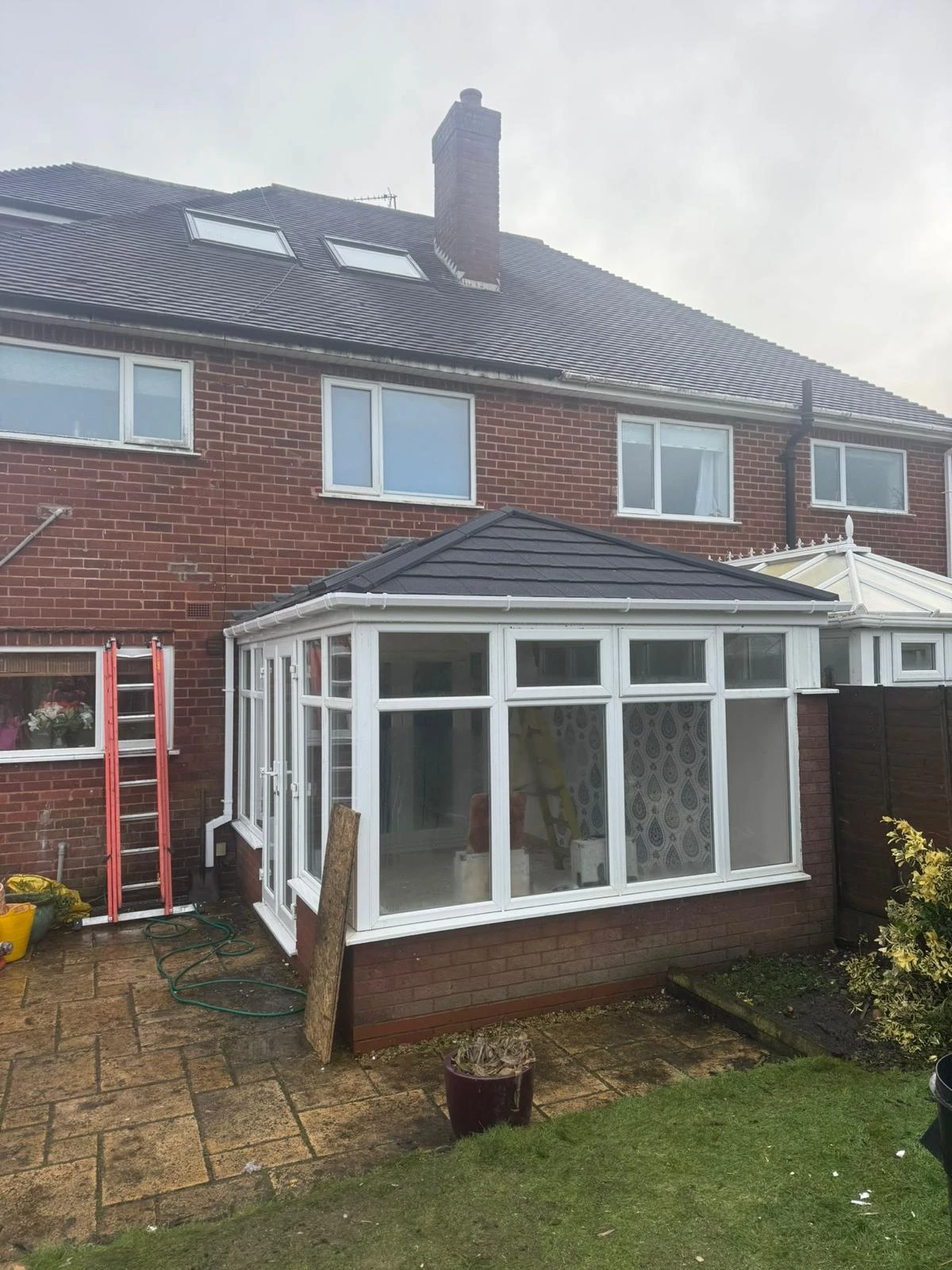 A brick house with a newly constructed conservatory, ladder, and pots in the garden. Grass and patio stones visible.