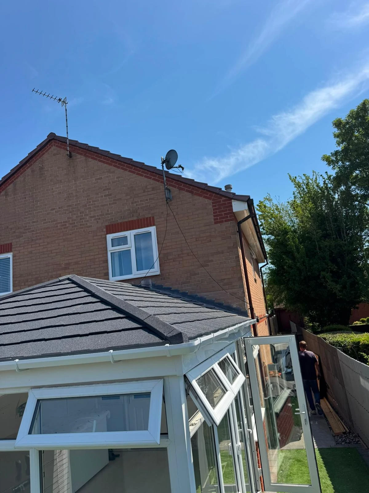 A brick house with a satellite dish and open conservatory door under a clear blue sky with scattered clouds.