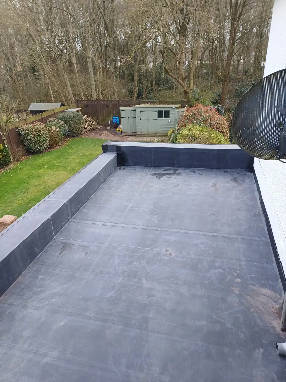View from a rooftop terrace overlooking a green yard with bushes, a shed, and trees in the background.