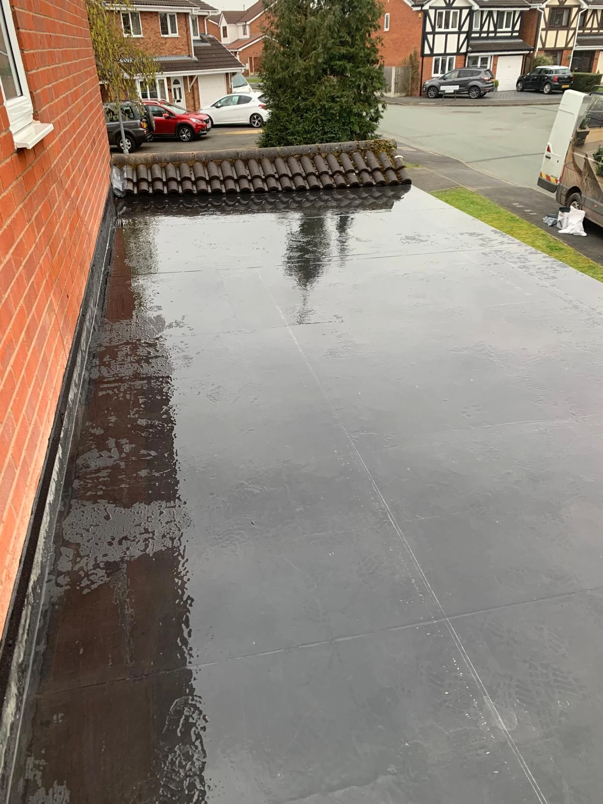 Flat roof showing a smooth, wet surface with a row of tile roofing nearby and residential houses in the background.