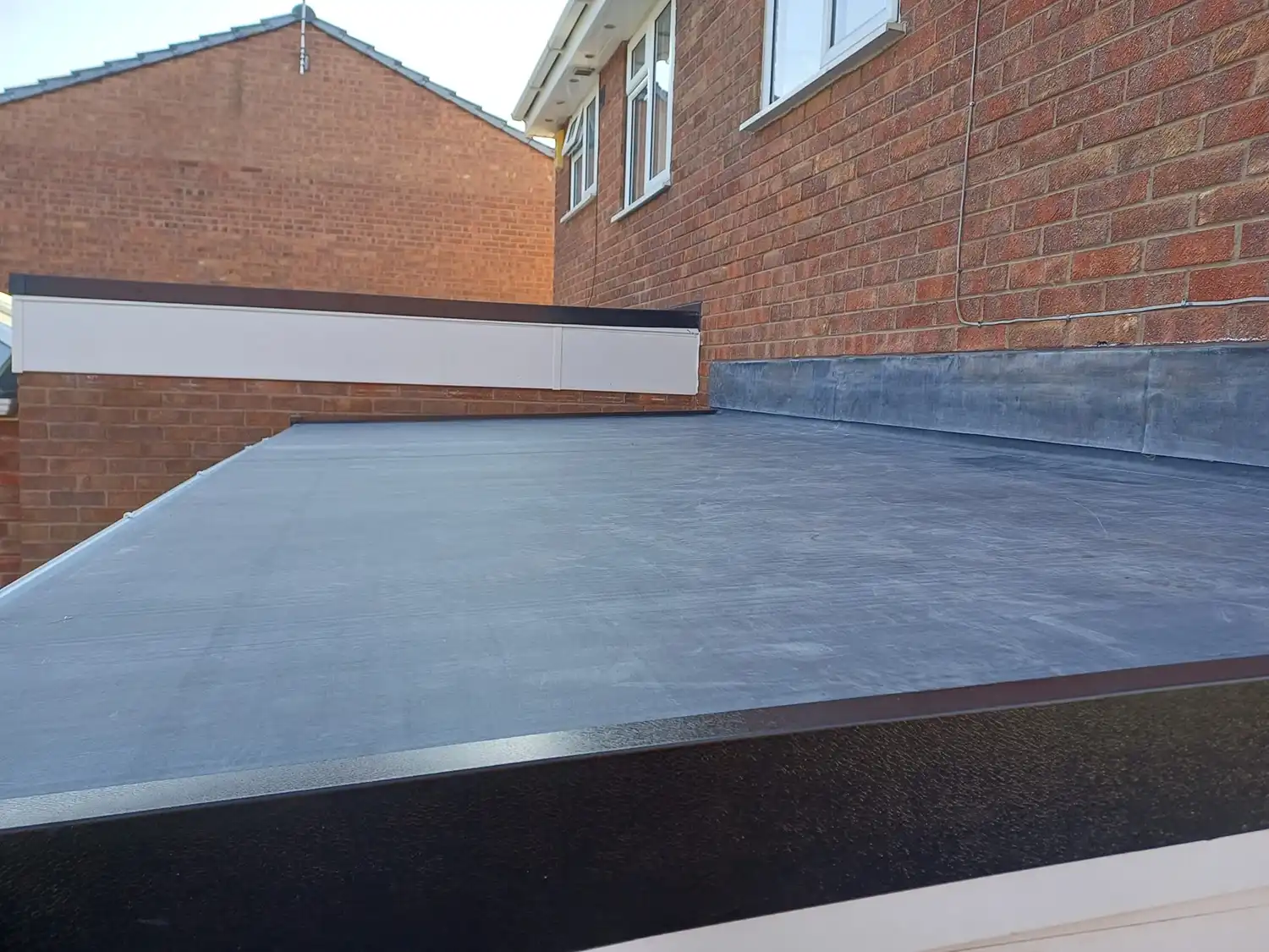 Flat roof with a smooth surface seen from a low angle, brick wall in the background, and a portion of a house visible.