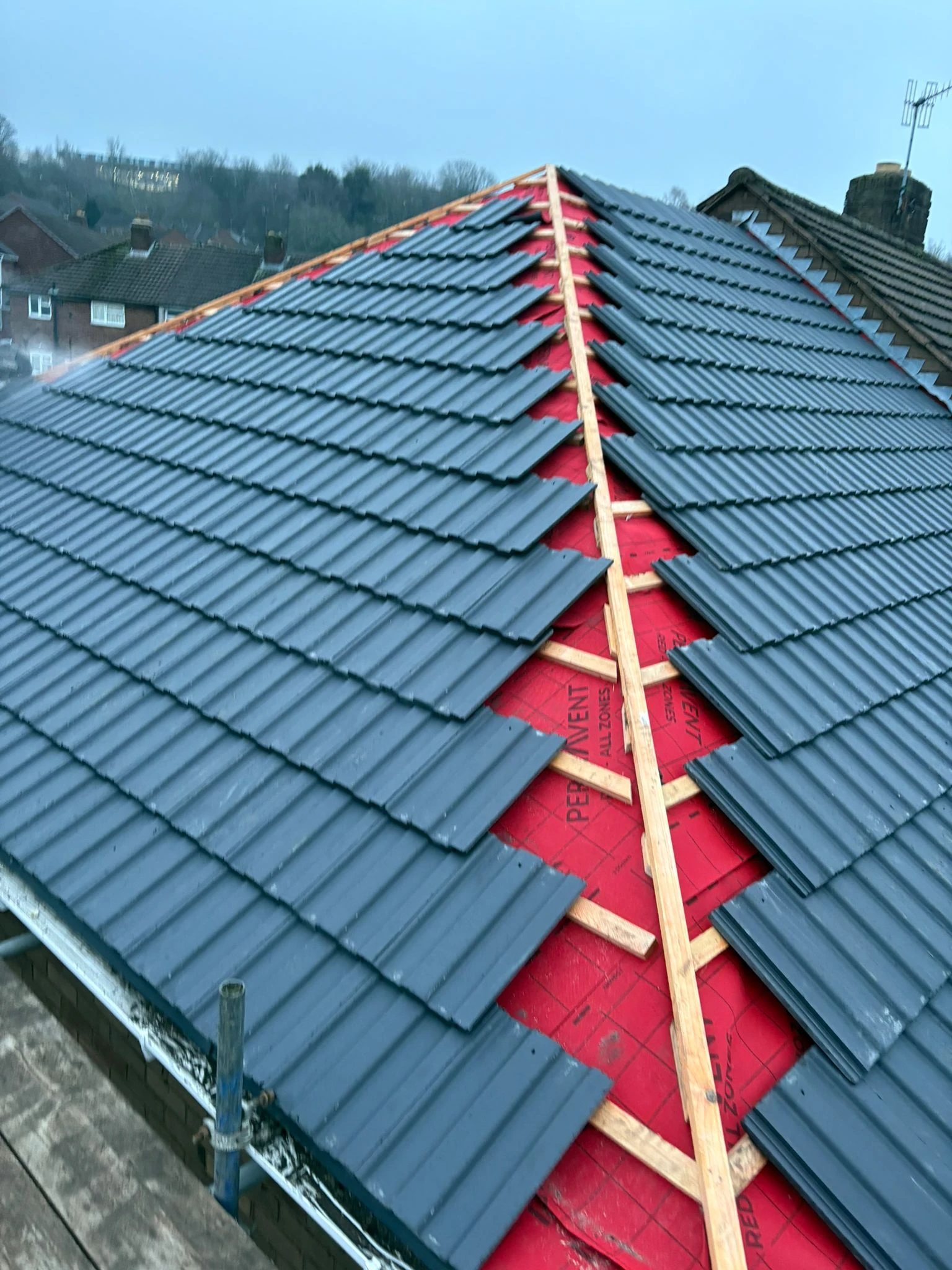 Newly installed dark grey roof tiles on a sloped roof, with red underlayment visible between tiles and wooden supports along the ridge.