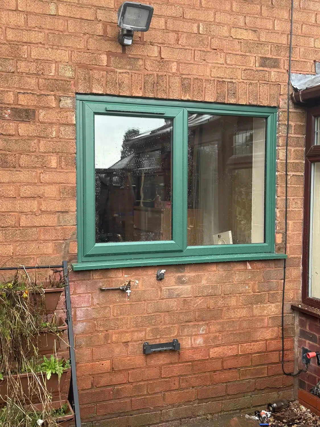 A green-framed window on a brick wall, with a light above it and planters beneath. Some outdoor clutter is visible.