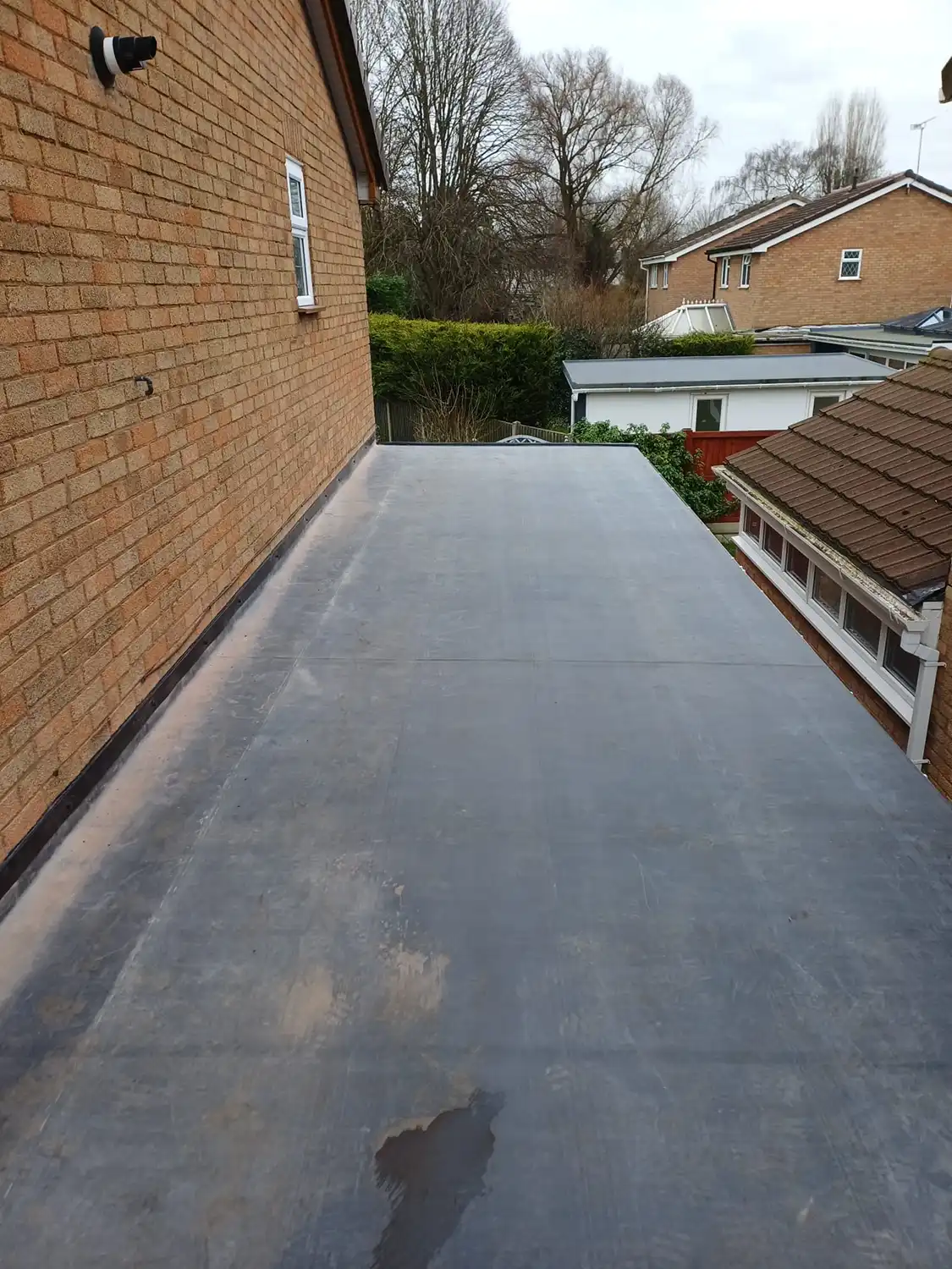 Flat roof view with a textured surface, adjacent to a brick wall and surrounding greenery, under a cloudy sky.